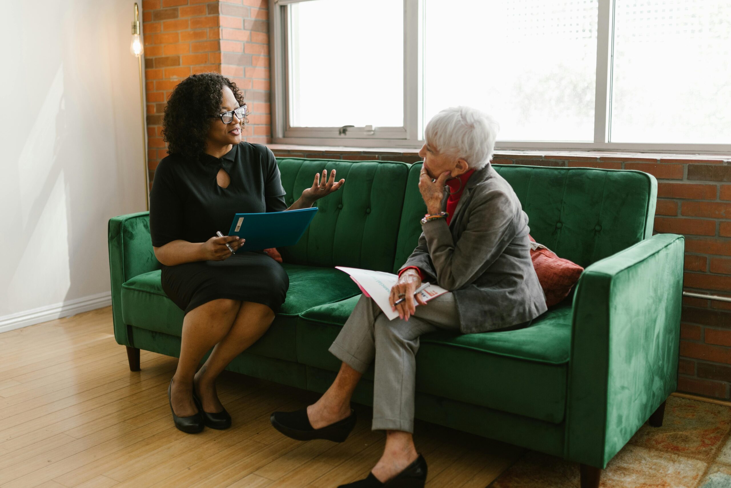 Two women engaged in a professional conversation sitting on a green sofa in a modern office setting.